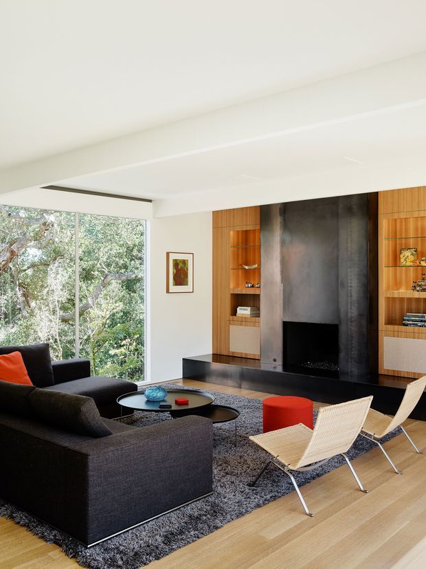 Bright contemporary living room with floor-to-ceiling glazing overlooking trees, a large dark sectional sofa, low round coffee tables, two woven lounge chairs, and a black fireplace feature wall integrated with warm timber display shelving and cabinetry. Light timber flooring and a textured charcoal rug create contrast against the white ceiling and walls.