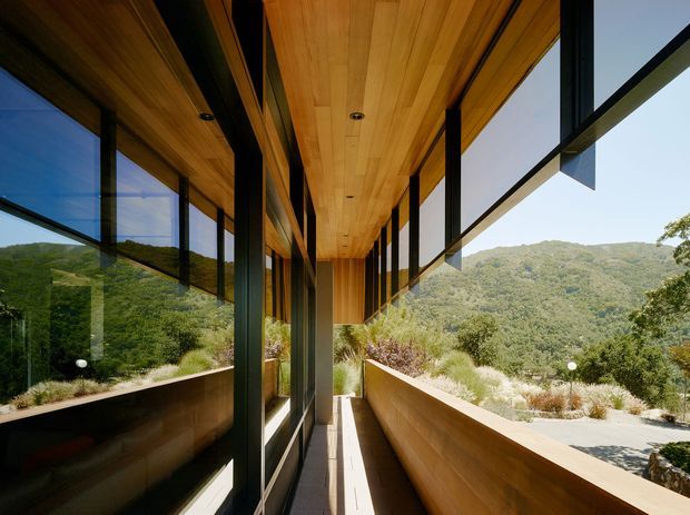 A narrow covered balcony or external walkway runs along a contemporary building, framed by extensive black-framed glazing and warm timber-lined soffits and walls. A timber balustrade and built-in planter edge border the walkway, with wooded hills visible beyond. The design emphasizes strong horizontal and vertical lines, deep overhangs, and a seamless connection to the surrounding landscape.