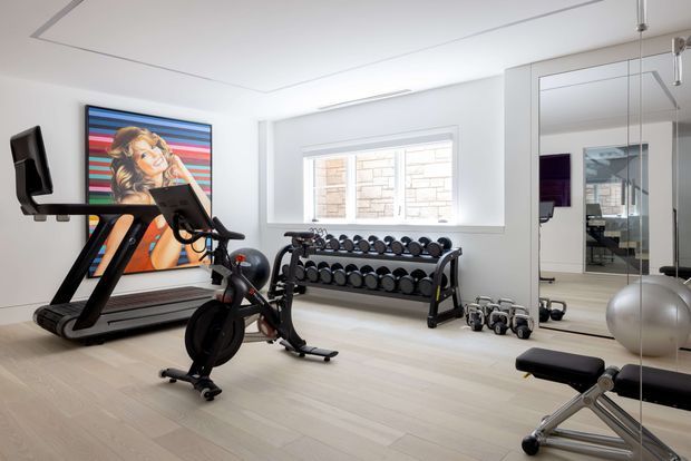 Bright, contemporary home gym with white walls, pale timber flooring, a treadmill, stationary bike, dumbbell rack, free weights, exercise bench and large mirrored wall. A colourful framed artwork adds a bold accent, while a window provides natural light. The mirror reflects additional gym equipment and a workspace beyond.