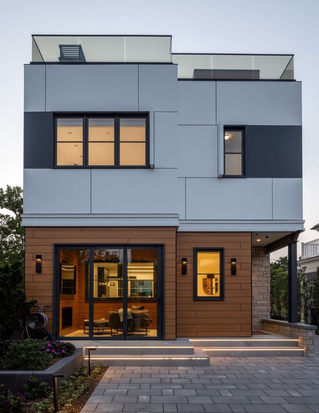 A contemporary multi-storey residential facade photographed from the front at dusk. The building combines smooth pale grey upper-level cladding with dark charcoal panels, warm horizontal timber-look cladding at ground level, extensive dark-framed glazing, rooftop terraces with clear glass balustrades, and a covered entry supported by a stone-clad pier. Broad grey paver steps and integrated linear lighting lead to the entrance, with landscaped planting along the edges.