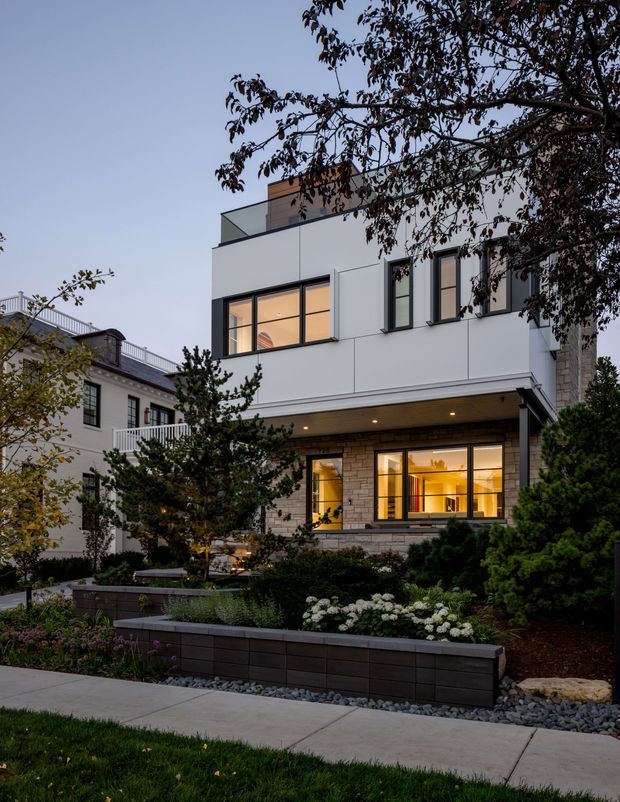 Evening exterior view of a contemporary multi-storey urban residence with a clean white facade, large dark-framed windows, a projecting upper level, rooftop terrace railing, and a warm stone-clad lower storey. The home is set behind a landscaped front garden with raised dark retaining walls, shrubs, flowers, and a paved footpath.