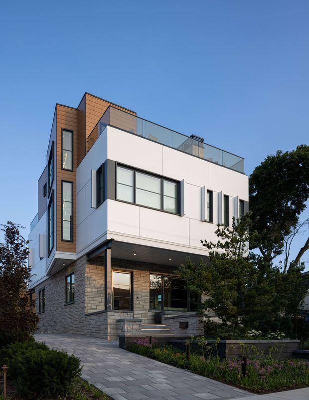 Exterior photograph of a contemporary multi-storey urban residence with a stepped, flat-roofed form. The facade combines smooth white wall panels, warm horizontal timber cladding, grey stone masonry at the lower level, dark-framed glazing, and a rooftop terrace enclosed by a glass balustrade. A paved path and landscaped garden lead toward the recessed entry and lower-level glazing.