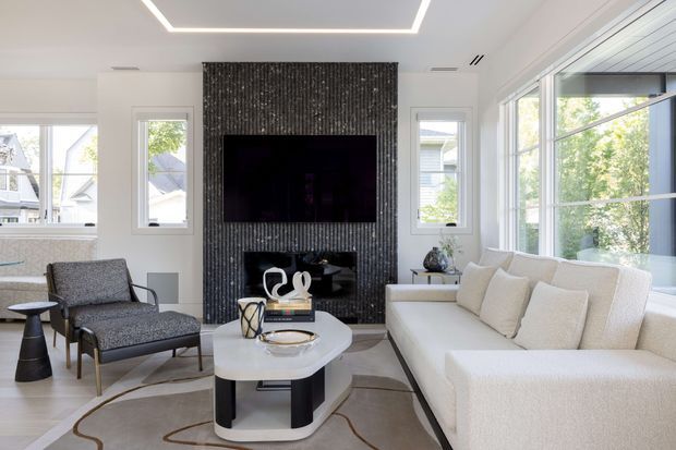 Bright contemporary living room with a large cream sectional sofa, sculptural white coffee table, patterned lounge chair, and wall-mounted television set into a dramatic dark textured feature wall. Large windows provide abundant natural light, while recessed linear ceiling lighting and pale flooring create an airy, refined interior.