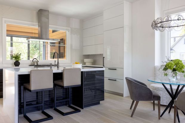 Bright contemporary kitchen and dining area with a central island, dark textured cabinetry, white full-height storage, pale wood-look flooring, and integrated stainless-steel appliances. Two upholstered stools sit at the island, while a small round glass dining table with upholstered chairs is positioned beside large windows. A modern sculptural pendant hangs above the dining area, and glazing provides views into an adjacent timber-lined space and outdoors.