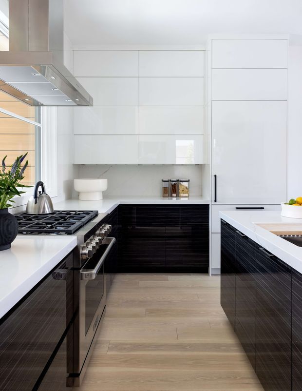 Bright, compact contemporary kitchen with a galley-style layout and central island. The room features glossy white full-height cabinetry, dark ribbed wood-look lower cabinetry, white benchtops, integrated appliances, a stainless-steel rangehood and cooktop, pale timber flooring, and large windows providing natural light.