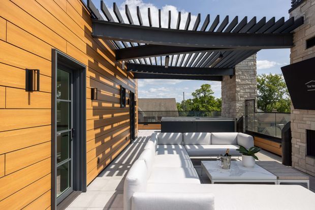 A bright contemporary rooftop or elevated outdoor lounge terrace with a white sectional sofa, low coffee table, built-in dark bar or counter, and a substantial stone-clad fireplace or wall. Warm horizontal timber-clad walls frame the space, while a dark steel pergola creates strong linear shadows across the pale terrace floor and furniture. Glass balustrades open views toward neighbouring rooftops and trees.