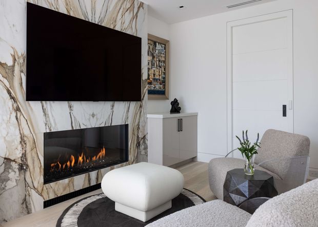 Contemporary living room centered on a dramatic bookmatched marble feature wall incorporating a wall-mounted television and linear built-in fireplace. The room includes a sculptural white ottoman, textured neutral lounge seating, a dark geometric side table, a compact grey built-in cabinet, and understated decorative accents. White walls, concealed ceiling services, and clean-lined doors reinforce the refined modern interior.