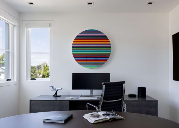 A clean, contemporary home office with a dark built-in desk spanning the wall, a central monitor, black ergonomic office chair, and compact desk accessories. White walls and ceiling, recessed downlights, and tall white-framed windows create a bright setting. A large circular multicoloured striped artwork provides a bold focal point above the desk, while an open book and closed laptop sit on the foreground table.