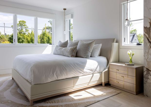 Bright, refined bedroom with a large upholstered bed dressed in white bedding and layered neutral pillows. The room features abundant natural light from multiple white-framed windows, a light timber bedside chest with drawer fronts, a slim bedside lamp, pale flooring, and a neutral patterned area rug. A marble-look vertical wall surface is visible at the right edge.
