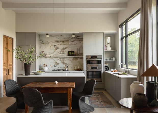 A refined open-plan kitchen and dining area with a long island, integrated appliances, pale grey cabinetry, and a dramatic veined stone backsplash. A timber dining table with woven dark chairs occupies the foreground, while large black-framed windows with full-length curtains bring in natural light. The room has a warm, sophisticated contemporary character with neutral finishes and timber accents.
