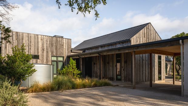 Exterior view of a low-rise contemporary building composed of weathered vertical timber cladding, with a prominent pitched gable roof and an attached flat-roofed carport or covered entry. The design uses simple rectangular volumes, dark metal roofing, timber posts and beams, and landscaped planting around a gravel-paved forecourt. A cylindrical water tank is visible beside the building.