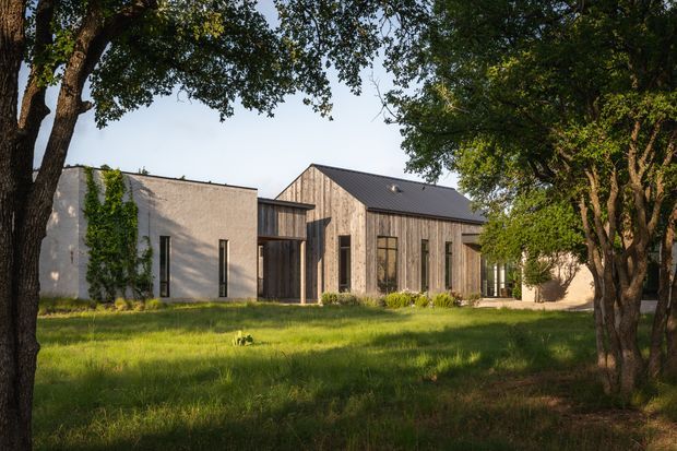 Exterior view of a contemporary single-storey residence set within a leafy landscaped garden. The building combines a simple pale rendered or masonry wing with a taller timber-clad gabled volume, dark pitched metal roofing, tall narrow windows, and a recessed glazed entry connection. Mature trees frame the foreground and cast dappled shade across the lawn.
