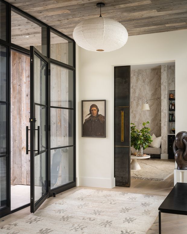 Refined residential entryway with a large pale patterned rug, black-framed glazed steel-style doors, and a warm reclaimed timber ceiling. A sculptural white pendant hangs centrally, while a portrait and dark built-in doorway cabinetry add character. Through the opening is a softly furnished living room with textured stone-look wall panels, shelving, a sofa, coffee table, and indoor greenery.