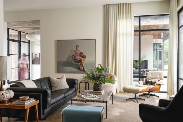 A refined contemporary living room with a black leather sectional sofa, sculptural lounge chairs, an ottoman, and a low rectangular coffee table. Full-height sheer curtains frame large black-framed windows and doors, opening visually toward a bright covered courtyard or outdoor area. The room features a neutral palette, a large framed artwork, layered soft furnishings, indoor greenery, and a glimpse of an adjacent dining or circulation space.