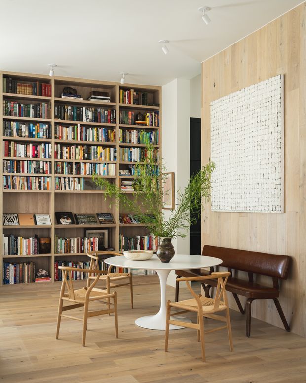 Bright, carefully styled dining and reading area with a large full-height timber built-in bookcase filled with books, a pale timber feature wall, and a white abstract artwork. A round white pedestal dining table is surrounded by timber armchairs, with a dark leather bench against the wall. Pale timber flooring, neutral walls, indoor plants, and ceiling-mounted spotlights create a warm contemporary interior.