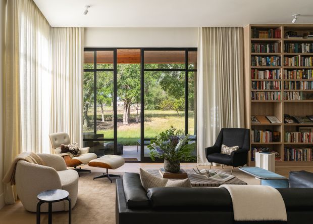 A refined contemporary living room with a large floor-to-ceiling glazed opening overlooking a leafy garden. The room features full-height cream curtains, a neutral upholstered armchair and sofa, a black leather sectional, a sculptural black lounge chair, a low coffee table, and extensive built-in timber book shelving along one wall. Natural light creates a calm, inviting atmosphere.