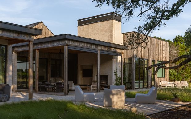 Exterior view of a contemporary single-storey residence with weathered timber cladding, broad covered outdoor living areas, large glazed openings, and a tall central chimney. The landscaped foreground includes a paved terrace with outdoor lounge furniture, while the building is set among mature trees.
