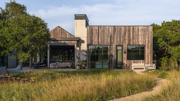 Single-storey contemporary home exterior set within a landscaped garden. The facade combines extensive vertical weathered timber cladding with dark-framed glazing, a rendered chimney, and a pitched timber-roofed section. Outdoor lounge furniture sits beneath a covered glazed porch, while ornamental grasses and mature planting occupy the foreground.