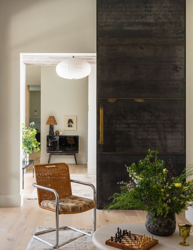 Refined interior view through a wide doorway, featuring pale timber flooring, soft beige walls, and a large dark aged timber or metal sliding door with a long brass handle in the foreground. A vintage woven lounge chair, mid-century console, framed artwork, table lamp, pendant light, chessboard, and a large floral arrangement create a warm, layered living space. The doorway and room beyond suggest an adjoining living room or sitting area.