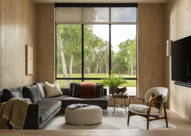 A warm, compact living room with full-height black-framed glazing overlooking a leafy garden. The room is enclosed in light timber wall panelling and flooring, with a dark sectional sofa, rounded ottoman, wood-framed lounge chair, small side tables, a potted plant, and a wall-mounted television. The composition is calm and contemporary, with abundant natural light and strong connection to the outdoors.