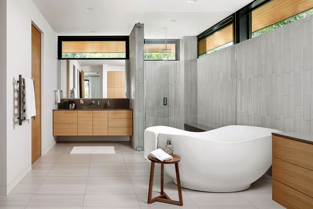 Contemporary bathroom featuring a freestanding white sculptural bathtub, double vanity with warm timber cabinetry and dark benchtop, large mirror, grey vertically patterned wall tiles, light floor tiles, and high clerestory windows with timber blinds. Black-framed glazing and black accents provide contrast in the bright, spacious room.