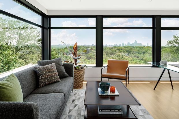 A bright, contemporary living room with expansive black-framed windows overlooking a leafy landscape and distant city skyline. The room features a large charcoal sectional sofa, geometric rug, low rectangular coffee table, tan leather armchair, minimalist desk or console, indoor plant, and pale timber flooring.