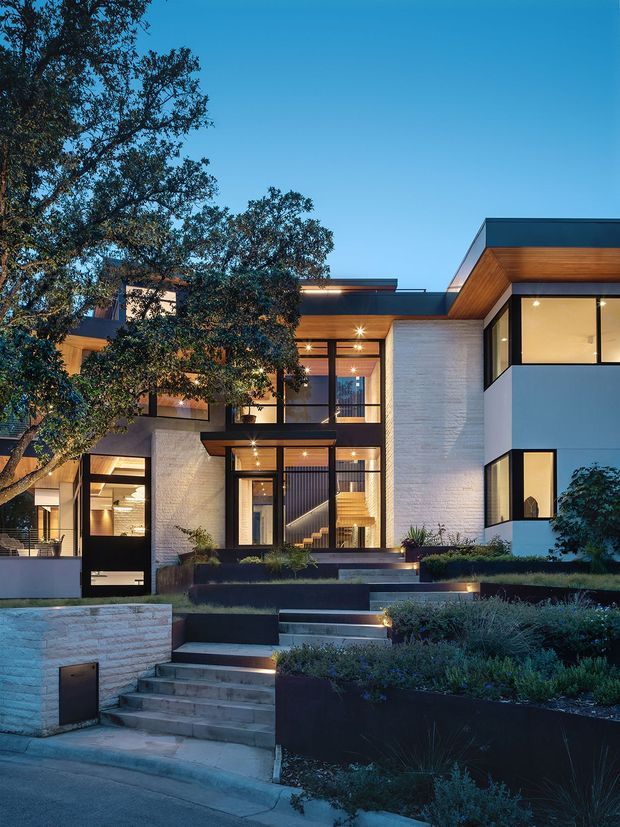 Twilight exterior photograph of a contemporary multi-level residence with strong geometric forms, extensive floor-to-ceiling glazing, white masonry walls, dark window frames, warm timber soffits, and layered landscaped terraces accessed by broad steps. The home has a flat-roofed composition and a welcoming illuminated entry courtyard.