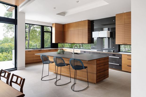 Contemporary open-plan kitchen with extensive warm timber cabinetry, a large central island with dark stone benchtop, integrated appliances, and three bar stools. Black-framed floor-to-ceiling glazing brings in abundant natural light and views of greenery. The space has a clean, refined, high-end feel with a recessed ceiling and modern stainless-steel and black fixtures.