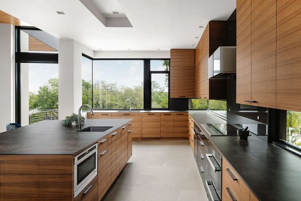 Contemporary open-plan kitchen with extensive warm timber cabinetry, dark stone benchtops, a central island with integrated oven and sink, black-framed floor-to-ceiling glazing, and a bright white ceiling with recessed linear skylight-like openings. The kitchen overlooks leafy greenery and has a clean, refined palette of timber, black, white, and grey.