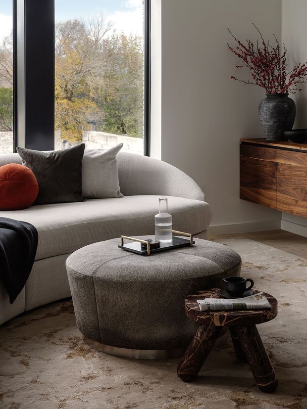 A refined contemporary living room featuring a large curved light-grey sofa, dark and pale cushions, a round upholstered ottoman, and a small sculptural timber side table. Floor-to-ceiling black-framed glazing overlooks trees and a low masonry wall. A floating timber media cabinet with a dark stone-like top and a textured black vase occupies the right wall. The room has neutral finishes, layered textiles, and soft natural light.