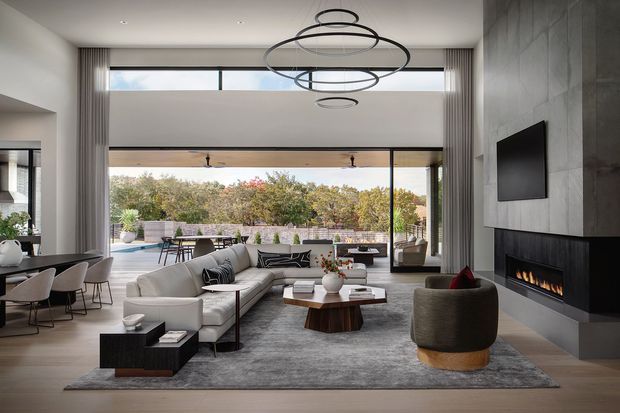 A sophisticated open-plan living room with a high ceiling, expansive floor-to-ceiling glazing, and seamless connection to an outdoor terrace and landscaped setting. The room features a large pale sectional sofa, sculptural lounge chairs, a timber coffee table, grey area rug, recessed dining area, a dramatic contemporary ring pendant, and a concrete-look feature wall incorporating a fireplace and wall-mounted television. The palette is neutral and refined, with abundant natural light and strong indoor-outdoor flow.