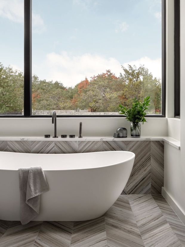 A bright, refined bathroom featuring a freestanding white bathtub positioned in front of large black-framed windows. The walls and floor are clad in chevron-patterned grey and taupe stone-look tiles, with a slim built-in ledge, wall-mounted bath fixtures, and a small plant on the sill. Trees and sky are visible beyond the glazing.