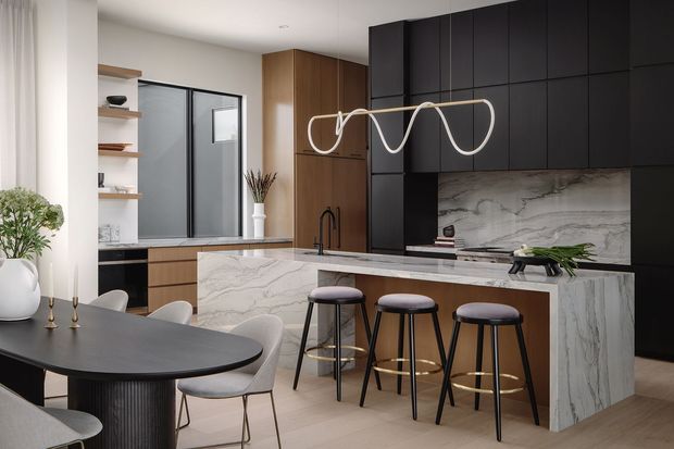 A contemporary kitchen and dining area with a large marble-look island, dark cabinetry, timber-toned joinery, integrated appliances, and three bar stools. A sculptural linear pendant hangs above the island, while a round dark dining table and upholstered chairs occupy the foreground. Full-height glazing and open shelving bring light into the refined, neutral-toned space.