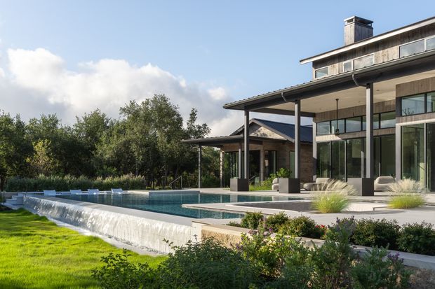 A contemporary multi-level residence overlooking a large landscaped swimming pool with an infinity-style overflow edge. The home features extensive floor-to-ceiling glazing, broad roof overhangs, dark slender structural posts, pale stone or concrete finishes, and a mix of timber-toned cladding and glass. The outdoor setting includes manicured lawn, planting, and a covered entertaining terrace under a clear blue sky.