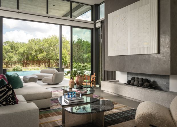 Contemporary living room with expansive black-framed floor-to-ceiling glazing overlooking a landscaped garden and pool. The room features a large light upholstered sectional sofa, sculptural lounge chairs, layered coffee tables, a patterned area rug, indoor plants, and a substantial dark textured fireplace wall with a long concrete or stone hearth and abstract wall artwork. The space is bright, refined, and closely connected to the outdoors.
