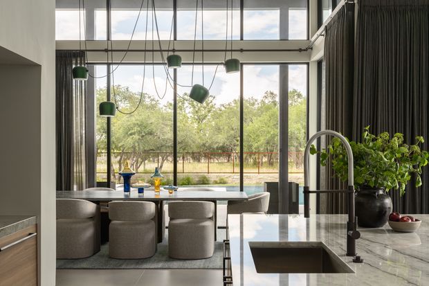 A sophisticated open-plan kitchen and dining area with a polished grey stone island, integrated sink, tall curved tap, dark cabinetry or wall cladding, and a large dining table beside floor-to-ceiling glazing. The dining space features rounded upholstered chairs and sculptural pendant lights, while greenery and an outdoor landscape are visible beyond the windows. The room has a high ceiling and a refined, contemporary feel.