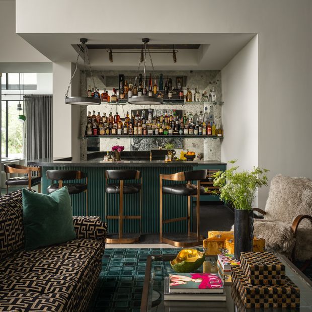A richly layered contemporary home bar and lounge area. The central built-in bar has dark vertically fluted cabinetry, a dark benchtop, brass-toned hardware, and mirrored or glass-backed shelving densely stocked with bottles. Three bar stools sit beneath suspended pendant lights. In the foreground are patterned upholstered lounge seating, a glass coffee table with books and decorative objects, and a textured armchair. A large window and curtains are visible to the left, while the room uses a sophisticated mix of deep green, charcoal, timber, brass, and warm neutral tones.