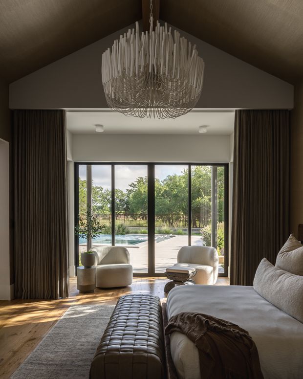 A refined bedroom with a large upholstered bed, sculptural tufted bench, two rounded lounge chairs, and a dramatic suspended chandelier. Full-height dark curtains frame expansive black-framed glazing that opens the room toward a landscaped patio and swimming pool. Warm timber flooring, neutral upholstery, and soft natural light create a luxurious, serene atmosphere.