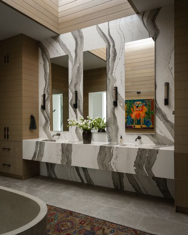 A sophisticated bathroom featuring a wide floating double vanity with integrated basins, large mirrors, and dramatic grey-veined white stone across the splashback, wall panels, and vanity front. Warm horizontal timber wall and ceiling panelling contrast with the stone. Black wall sconces, dark cabinetry, a colourful framed artwork, a freestanding curved bathtub edge, and a patterned rug complete the refined interior.