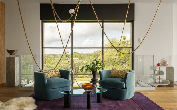 A refined contemporary living room with two deep teal armchairs arranged around a glass coffee table on a purple rug. Floor-to-ceiling glazing provides expansive views of leafy landscape, while sculptural looping pendant cords, warm timber flooring, and curated decorative objects create a dramatic yet inviting interior.