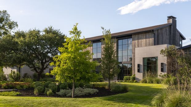 A large contemporary two-storey residence set within a carefully landscaped garden. The facade combines dark vertical timber cladding, smooth light stucco or concrete-like wall planes, extensive black-framed floor-to-ceiling glazing, and a low pitched roof with a prominent chimney. Curved planting beds, mature trees, ornamental grasses, and a manicured lawn frame the building.