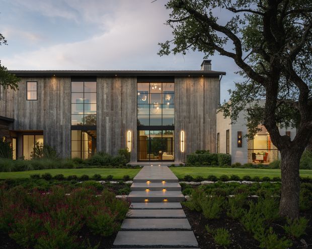 A contemporary two-storey residence viewed from the landscaped front garden at dusk. The facade combines vertically weathered timber cladding with pale rendered masonry, a low-pitched dark roof, and a tall central glazed entry void. A straight path with illuminated steps leads through layered planting to the entrance, while mature trees frame the composition.
