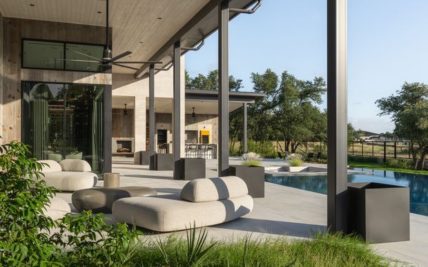 A luxurious contemporary indoor-outdoor living space with large floor-to-ceiling glazing opening onto a swimming pool and landscaped grounds. The covered terrace features sculptural neutral sofas, dark structural columns, a timber-lined ceiling, an outdoor ceiling fan, planters, and views through to a modern kitchen and fireplace area. The design strongly emphasizes seamless connection between interior and exterior.