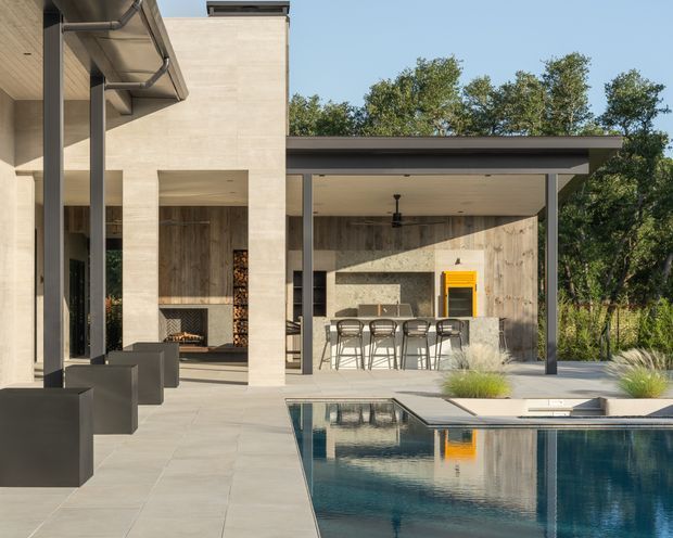 A contemporary residential outdoor entertaining area beside a rectangular swimming pool. The design features pale stone or concrete-clad walls, slim dark metal posts, a broad flat roof, large openings, an outdoor kitchen with barbecue and island seating, and a covered lounge zone. The pool and paving create strong reflections, while mature vegetation frames the background.
