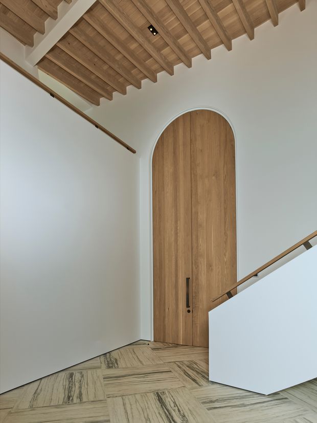 A refined interior stair landing featuring a very tall arched timber door set into smooth white walls. The ceiling has exposed timber rafters and boards, while the floor is laid in a distinctive patterned natural stone or marble with strong linear veining. A minimalist staircase with a white solid balustrade and slim timber handrail enters from the right.