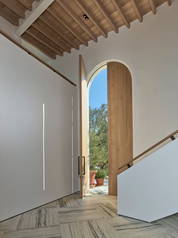 A refined residential entry hall viewed from inside, featuring a tall arched timber front door opened to a planted outdoor area. White plaster walls contrast with a dramatic exposed timber-rafter ceiling, while the floor is finished in large-format veined stone tiles. A simple contemporary stair with a white solid balustrade and timber handrail rises at right.
