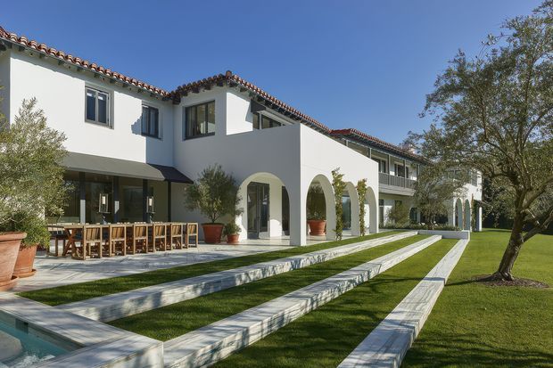 Exterior view of a large white stucco residence with terracotta tiled roofs, repeated arched openings, upper-level balconies, and expansive landscaped lawns. Long pale stone or concrete paths create strong linear bands across the grass, while a covered outdoor dining terrace with timber furniture sits beside the house. The architecture has a Mediterranean or Spanish-influenced character in a bright, formal garden setting.
