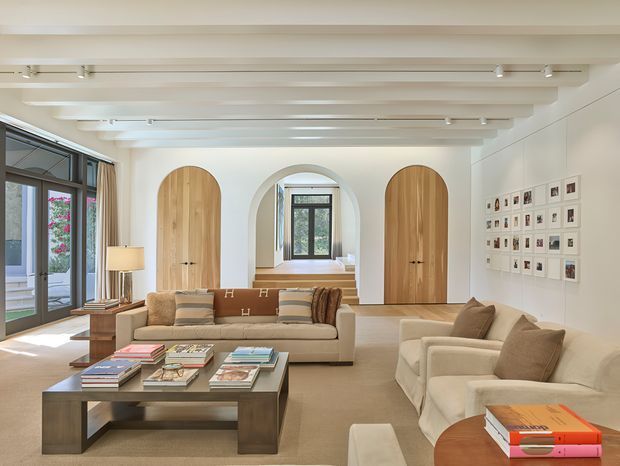 Bright, refined living room with a warm neutral palette, exposed white-painted ceiling beams, pale timber flooring, arched timber doors, and a central arched window opening. The room features a large upholstered sofa, two lounge chairs, a substantial dark coffee table with books, extensive glazing to the left, and a curated grid of framed artwork on the right wall.