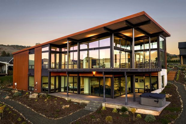Exterior architectural photograph of a contemporary multi-level house on a landscaped sloping site. The home features extensive floor-to-ceiling glazing, dark slender structural framing, warm timber wall cladding, projecting roof planes, elevated upper-level living spaces, and a covered lower terrace with outdoor seating. Hills and neighbouring buildings are visible in the background.