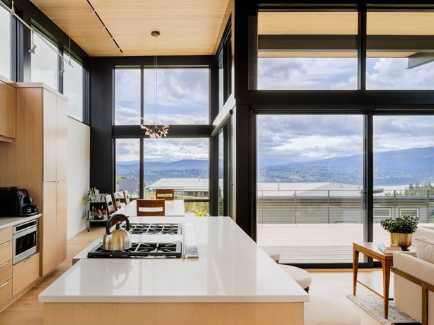 A bright, open-plan contemporary kitchen and living area with a large white island, integrated gas cooktop, light timber cabinetry, and expansive black-framed floor-to-ceiling glazing. The glazing opens to a deck or terrace with mountain and valley views. A timber-lined ceiling, black feature walls and frames, and warm natural light create a refined connection between the interior and landscape.